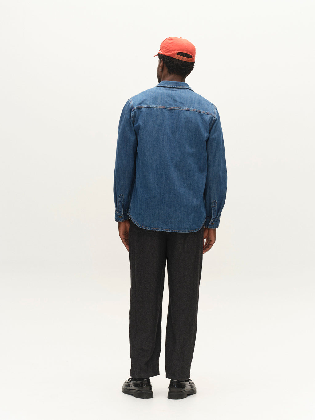 A person stands facing away, wearing a red cap, dark pants, and black shoes against a plain, light background. The highlight of their outfit is the Gabba GABsven Reg Wish LS Shirt in Mid Blue Denim—a versatile wardrobe essential.