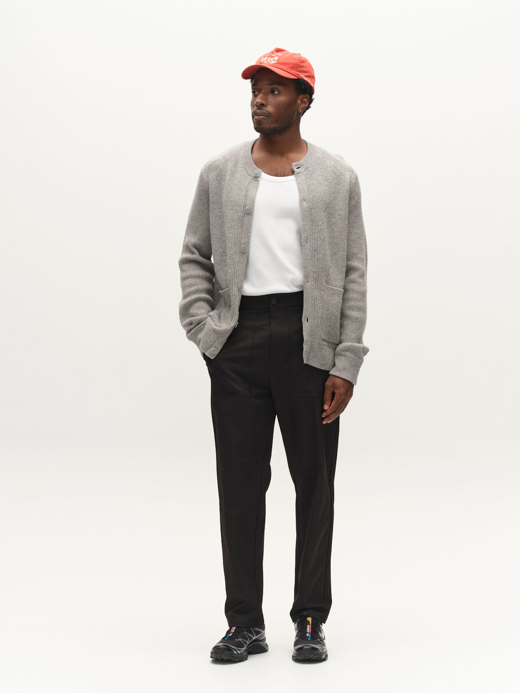 A man wearing a red cap, stylish Gabba GABremy Cardi Flash LS Knit in Grey Mel over a white T-shirt, paired with black pants and black shoes, stands against a plain white background with one hand in his pocket, glancing to the side.