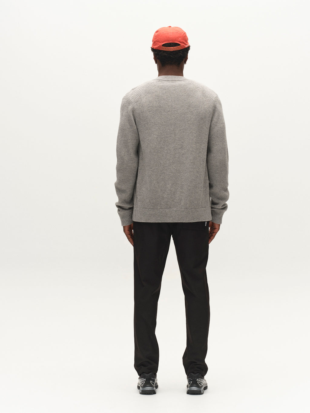 A person stands facing away from the camera, wearing the Gabba GABremy Cardi Flash LS Knit in Grey Mel, paired with black pants, black shoes, and a red cap, against a plain white background.