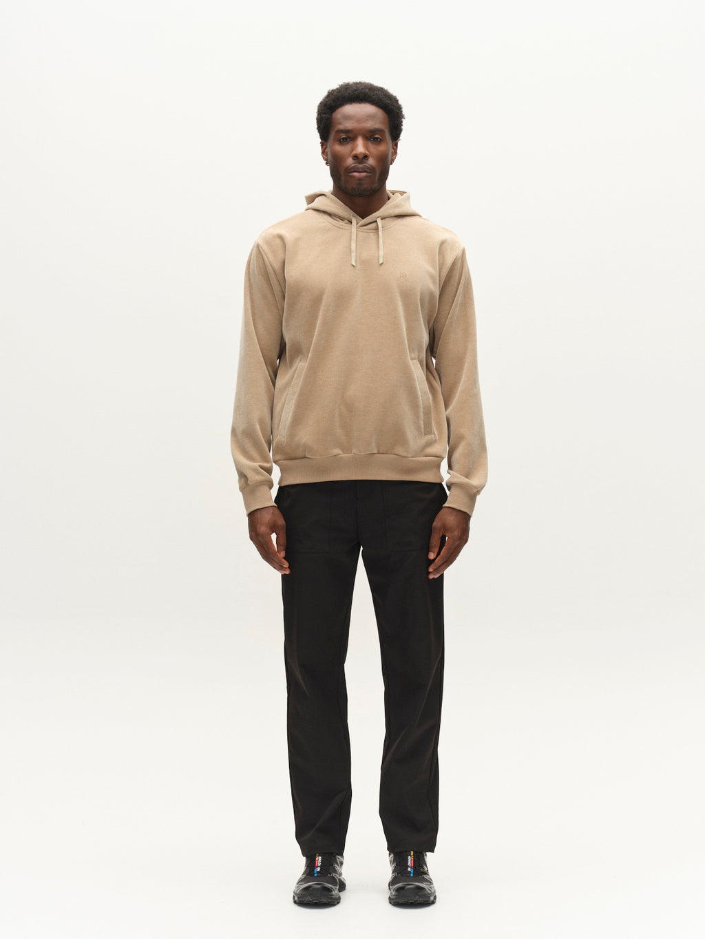 A man stands facing forward wearing the Gabba GABprimo Fune Hoodie LS Sweat in Sand Crocker, paired with black pants and black shoes against a plain white background.