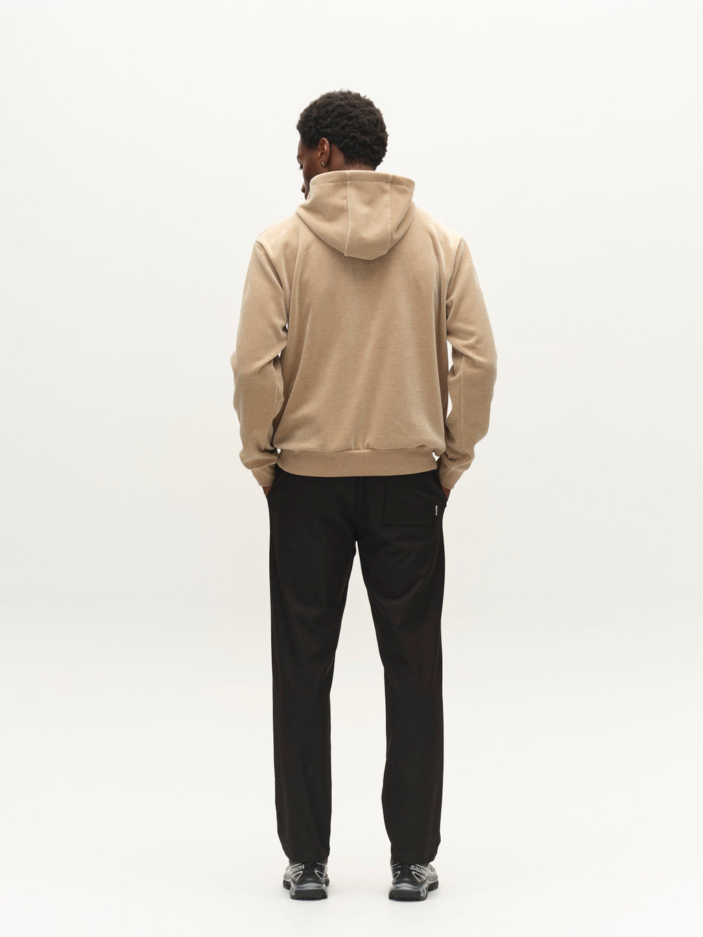A person stands facing away, wearing the Gabba GABprimo Fune Hoodie LS Sweat in Sand Crocker, paired with black pants and black sneakers, against a plain white background.