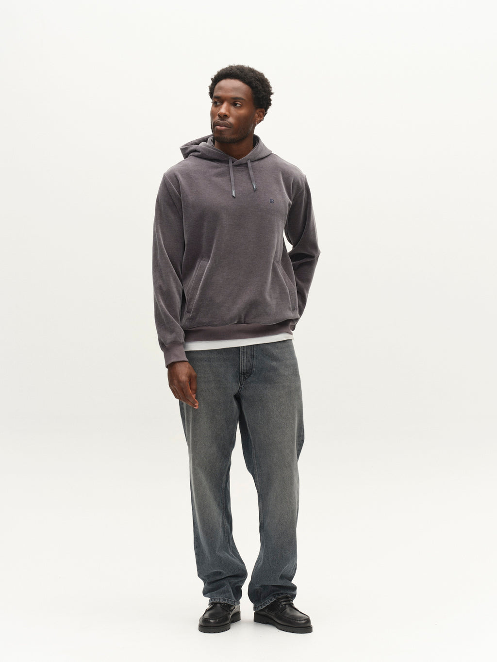 A man wearing the Gabba GABprimo Fune Hoodie LS Sweat in Turbulence Blue, paired with jeans and black shoes, stands facing slightly left against a plain white background, with his left hand tucked into his pocket. The regular fit of the hoodie enhances his relaxed yet stylish look.