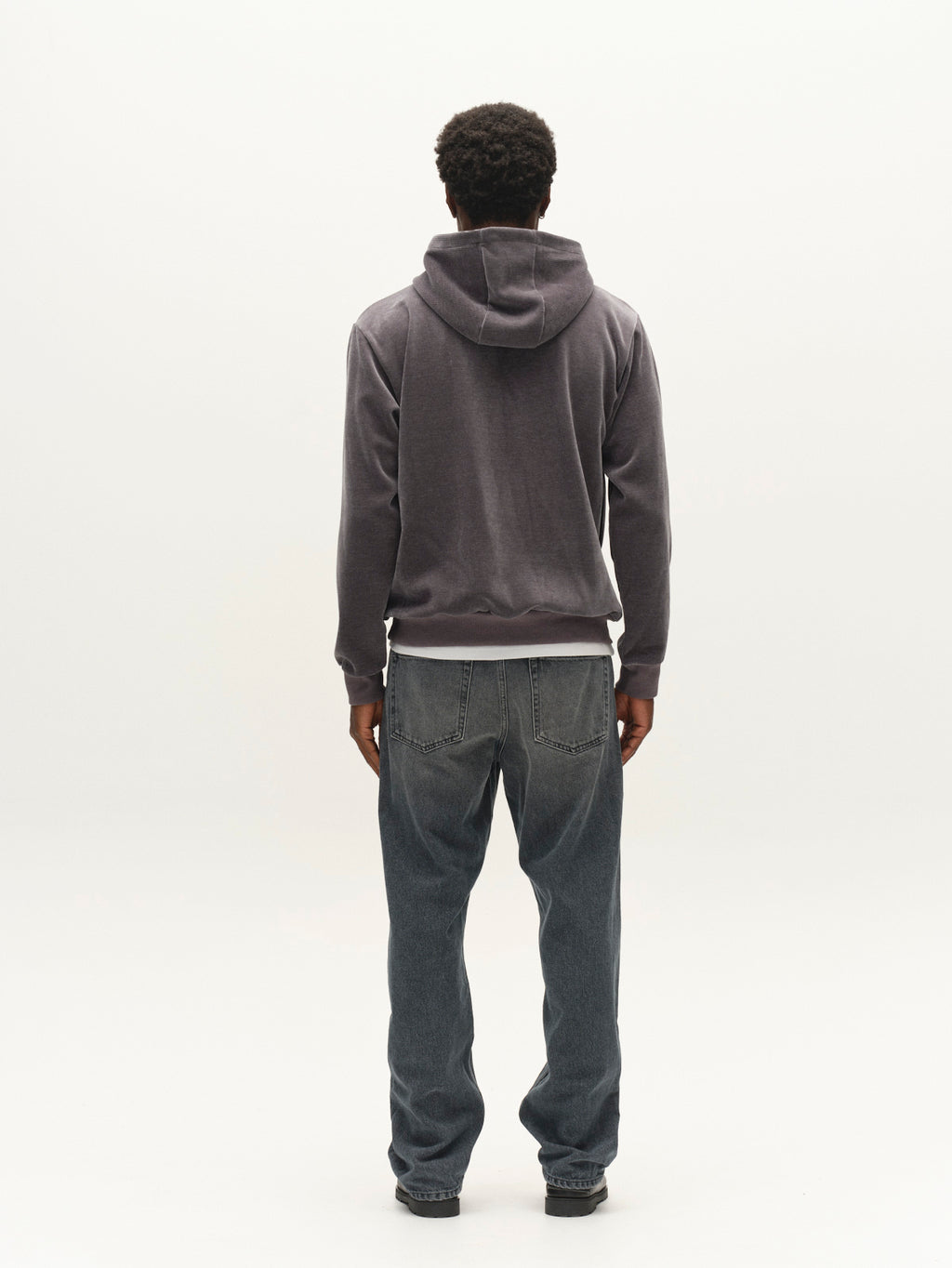 A person stands facing away, wearing the Gabba GABprimo Fune Hoodie LS Sweat in Turbulence Blue, paired with blue jeans and black shoes, against a plain white background.
