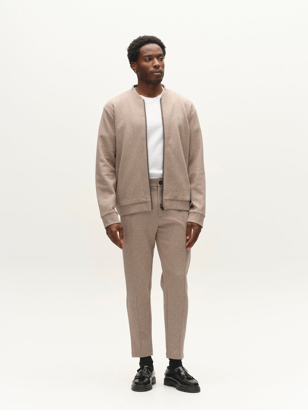 A man stands against a plain white background wearing the Gabba GABnik Camp Lock Sweat in Sand Mel., paired with matching beige pants, a white t-shirt, and black shoes—a contemporary showcase of modern outerwear.