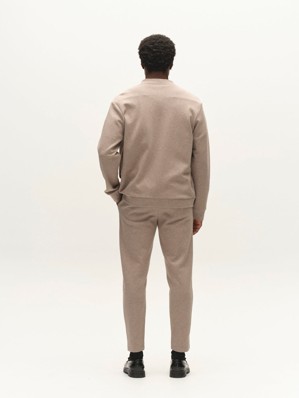 A person stands facing away from the camera, wearing the Gabba GABnik Camp Lock Sweat in Sand Mel—a matching light brown long-sleeve top and pants crafted from mid-weight interlock fabric—with black shoes, set against a plain white background.