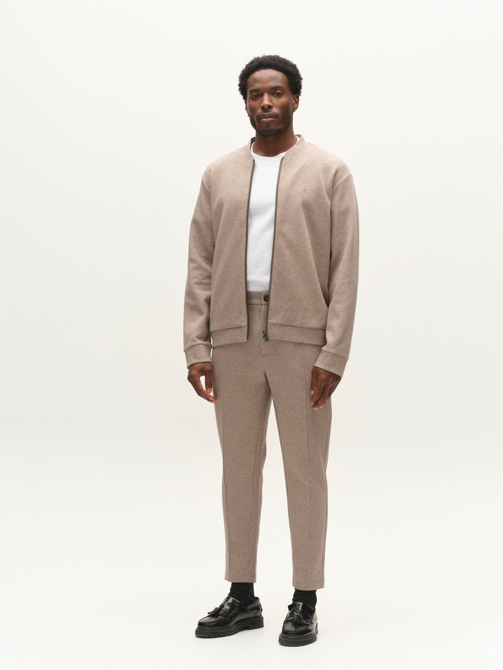A man stands facing forward against a plain white background, wearing a tan zip-up jacket, a white shirt, and black shoes. His pants are the Gabba GABmonza Cropped Tasca Lock Pant in Sand Mel, featuring a relaxed straight fit with cropped legs and crafted from mid-weight interlock fabric.