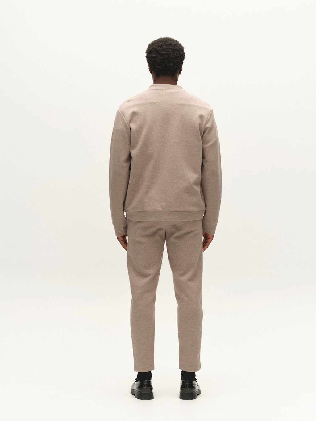 A person stands facing away from the camera, wearing the Gabba GABmonza Cropped Tasca Lock Pant in Sand Mel, featuring a relaxed straight fit and cropped leg, paired with a light brown jacket and black shoes against a plain white background.