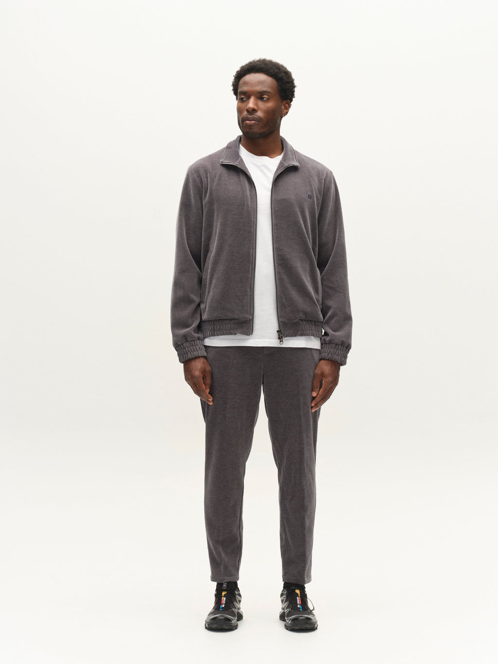 A man stands facing forward, wearing a gray zip-up jacket, a white T-shirt, black sneakers, and Gabba GABmonza Cropped Tasca Fune Pants in Turbulence Blue against a plain white background.