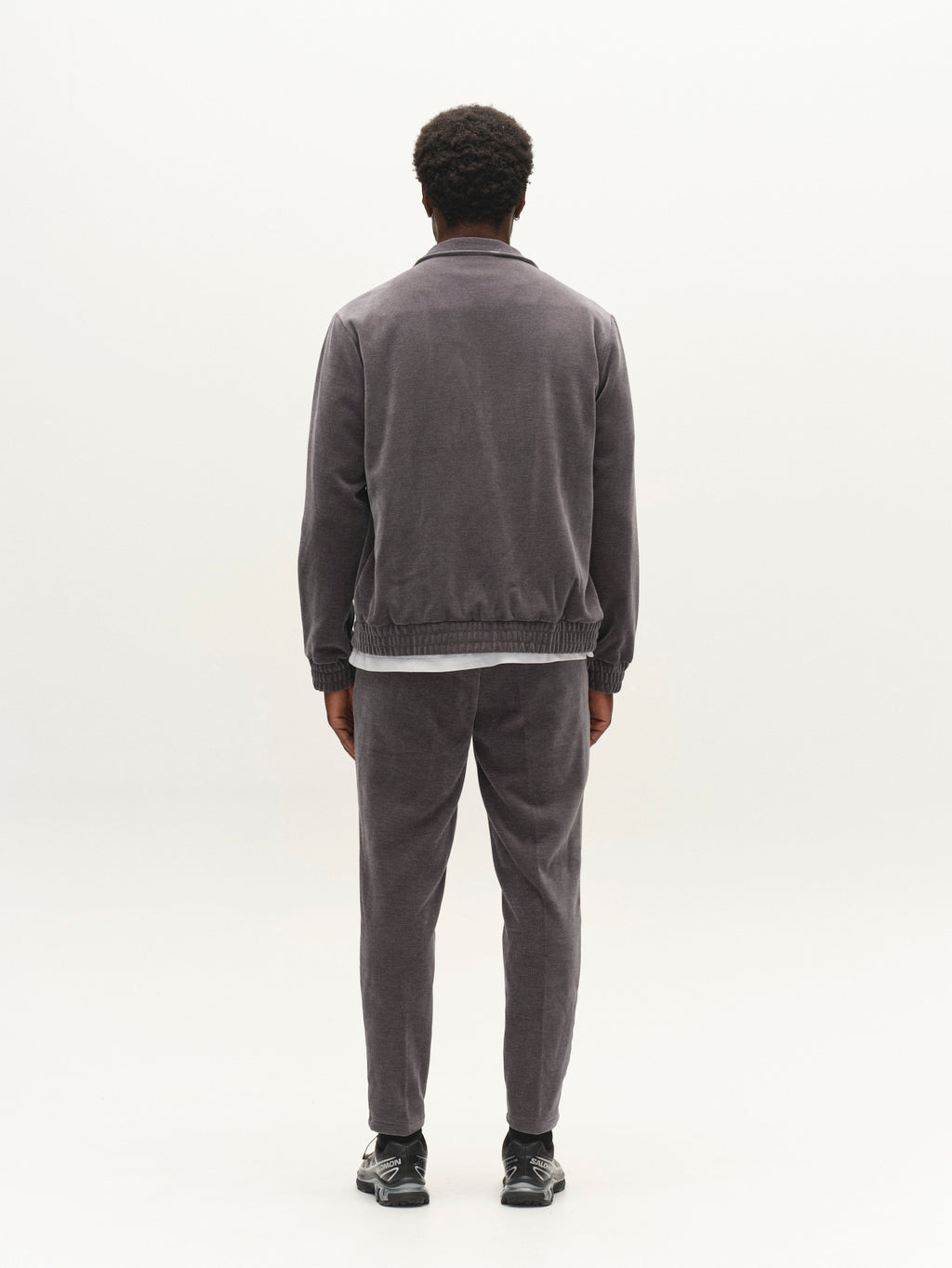 A person with short curly hair, dressed in a grey tracksuit and black shoes, stands facing away from the camera against a plain white background, showcasing the Gabba GABmonza Cropped Tasca Fune Pant in Turbulence Blue—a relaxed straight fit jersey corduroy style with an elastic waistband.