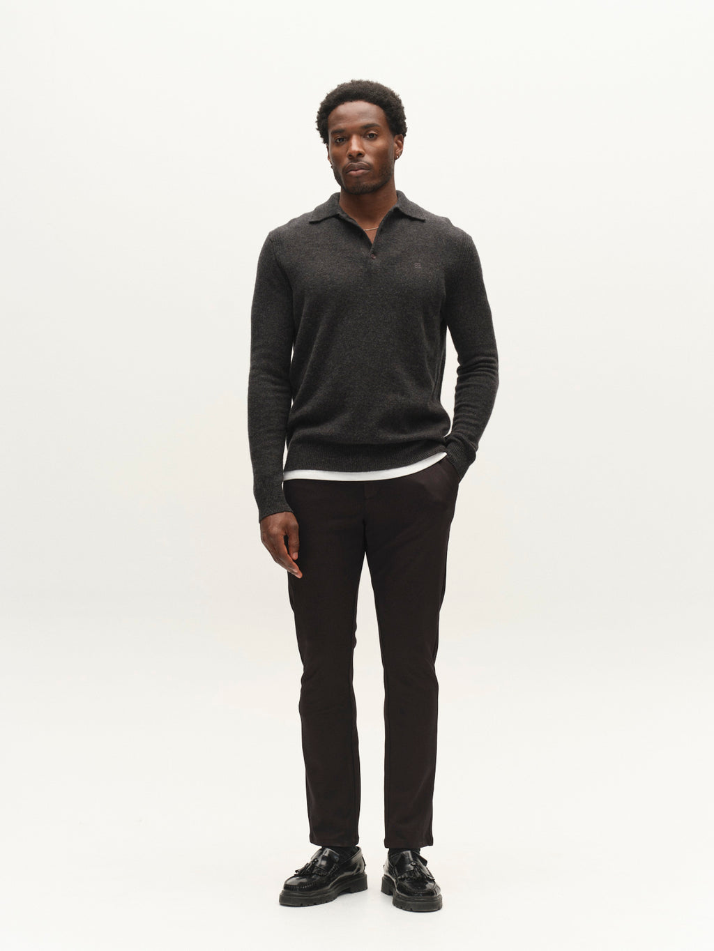 A man stands against a plain white background, wearing a dark sweater over a white shirt, black shoes, and the Gabba GABpaul Dotty Pant in Meteorite—a slim chino fit crafted from superstretch fabric—with one hand in his pocket.