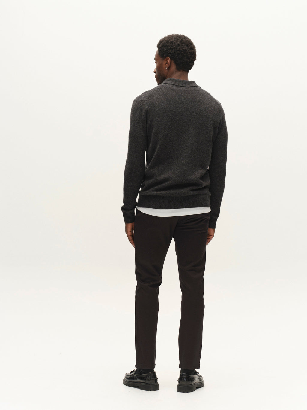 A man with short curly hair, dressed in a dark sweater, Gabba’s GABpaul Dotty Pant in Meteorite—a slim fit chino crafted from superstretch fabric—and black shoes stands facing away in a plain white studio setting.