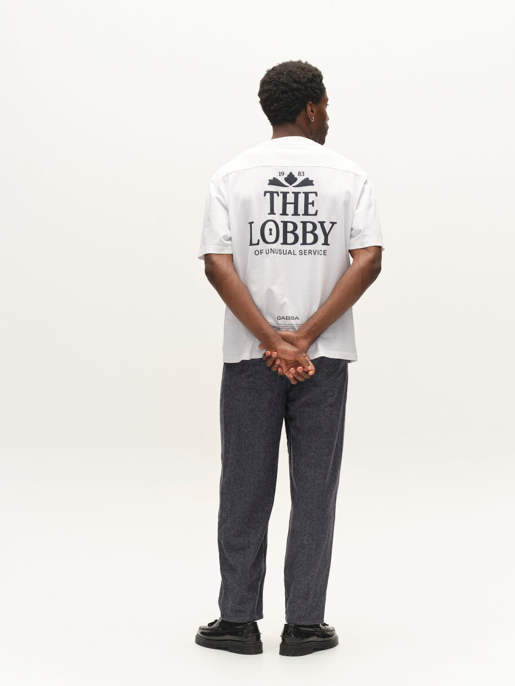 A person stands facing away with hands behind their back, wearing a white shirt that reads THE LOBBY OF UNUSUAL SERVICE and dark navy GABdavid Twilight Pants by Gabba in a contemporary chino style with a loose tapered fit, against a plain white background.