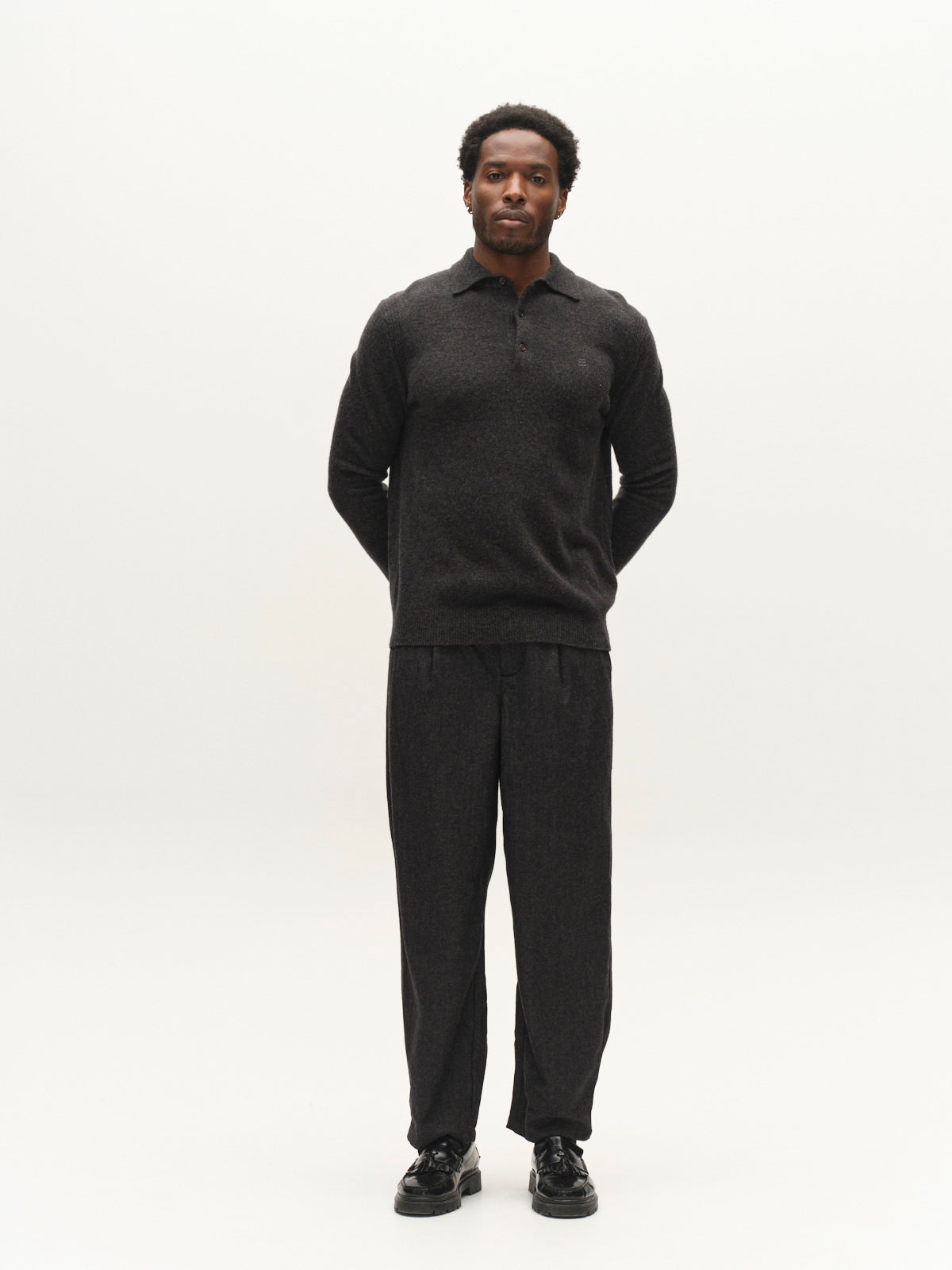 A man stands facing forward with his hands behind his back, wearing a dark long-sleeve shirt and the Gabba GABdavid Twilight Pant in Dark Grey, featuring a loose tapered fit, paired with black shoes against a plain light background.