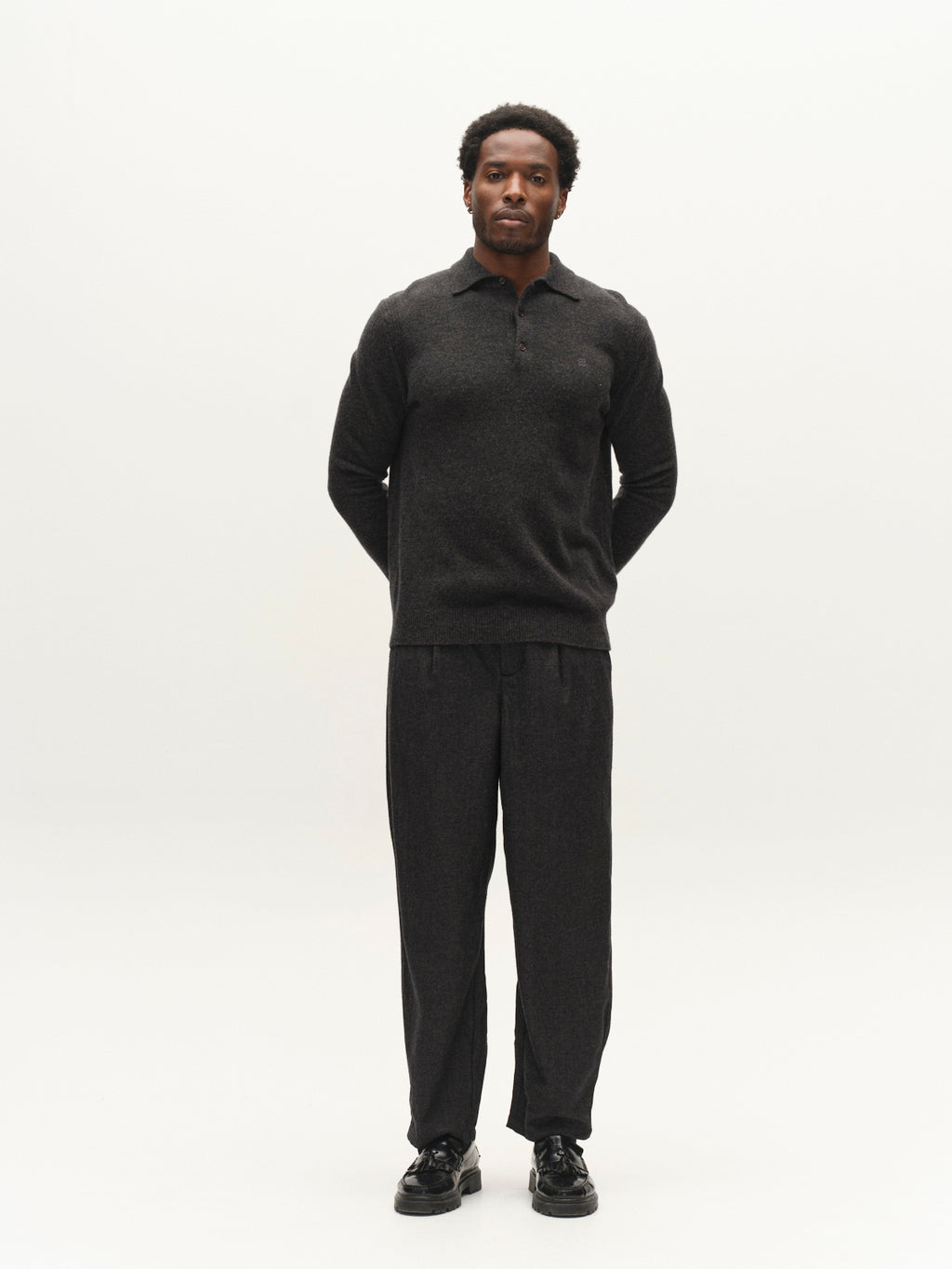 A man stands facing forward with his hands behind his back, wearing a dark long-sleeve shirt and the Gabba GABdavid Twilight Pant in Dark Grey, featuring a loose tapered fit, paired with black shoes against a plain light background.