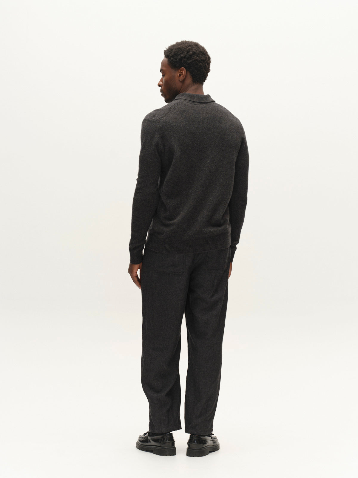 A man stands facing away, wearing a dark sweater, black shoes, and the Gabba GABdavid Twilight Pant in Dark Grey against a plain white background.