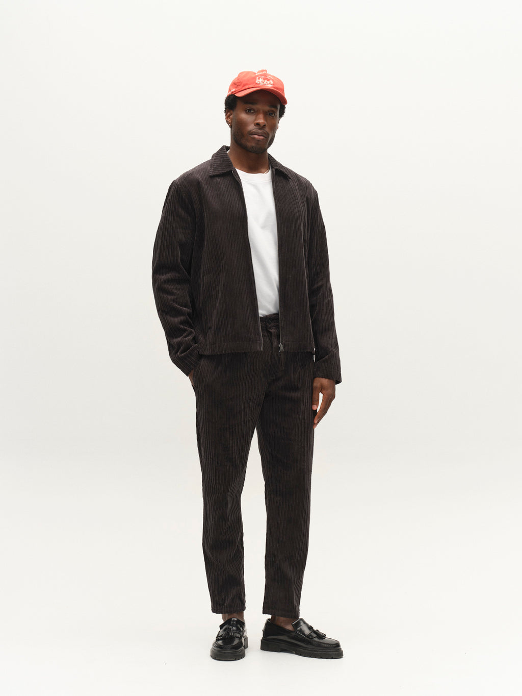 A man stands in a studio wearing the Gabba GABfirenze Cropped Riglia GOTS Pant in dark grey with a relaxed fit, paired with a matching corduroy jacket, white T-shirt, black shoes, and a red cap. The background is plain and light-colored.