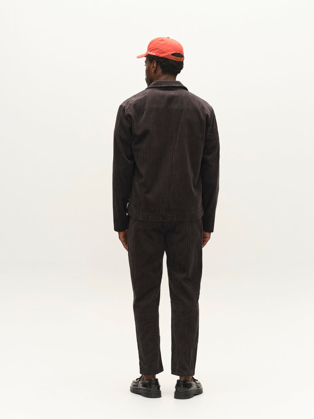 A man stands facing away against a plain white background, wearing a relaxed-fit dark organic cotton corduroy jacket, the Gabba GABfirenze Cropped Riglia GOTS Pant in Dark Grey, black shoes, and a red cap.