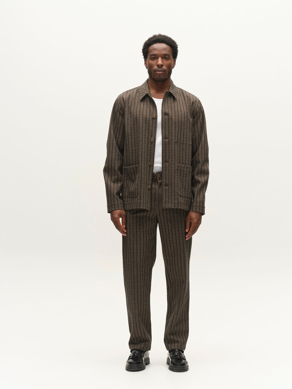 A man stands facing forward against a plain white background, wearing Gabba’s GABnapoli 19 Zapies Pant in Brown Slate styled with a matching pinstriped jacket over a white shirt and black shoes.
