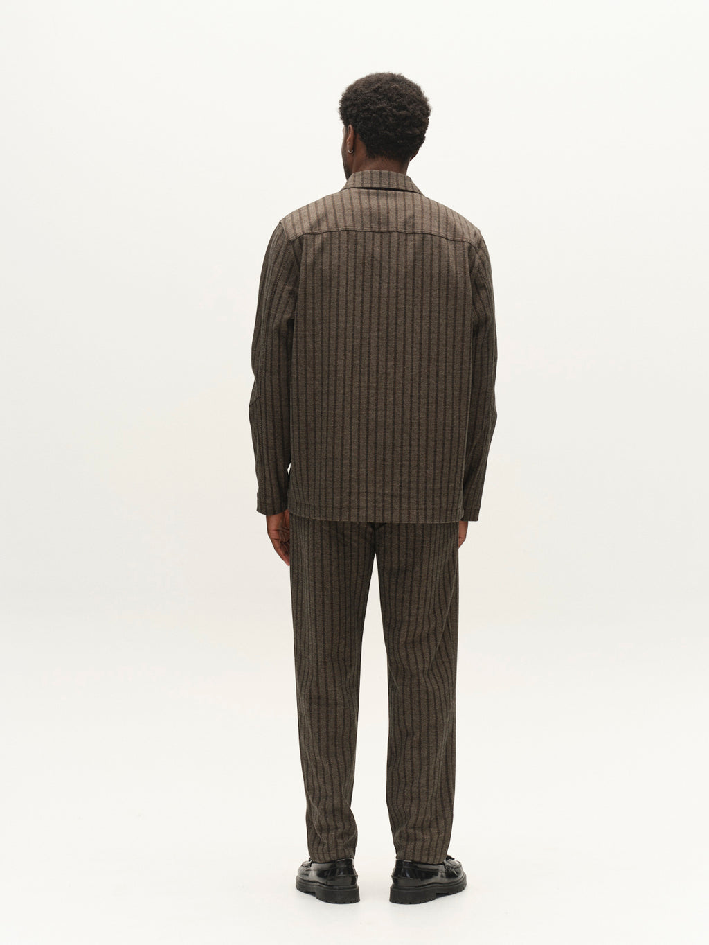 A person stands facing away, wearing the Gabba GABnapoli 19 Zapies Pant in Brown Slate, featuring a matching wool-look stripe stretch fabric with regular straight fit trousers and black shoes, on a plain white background.