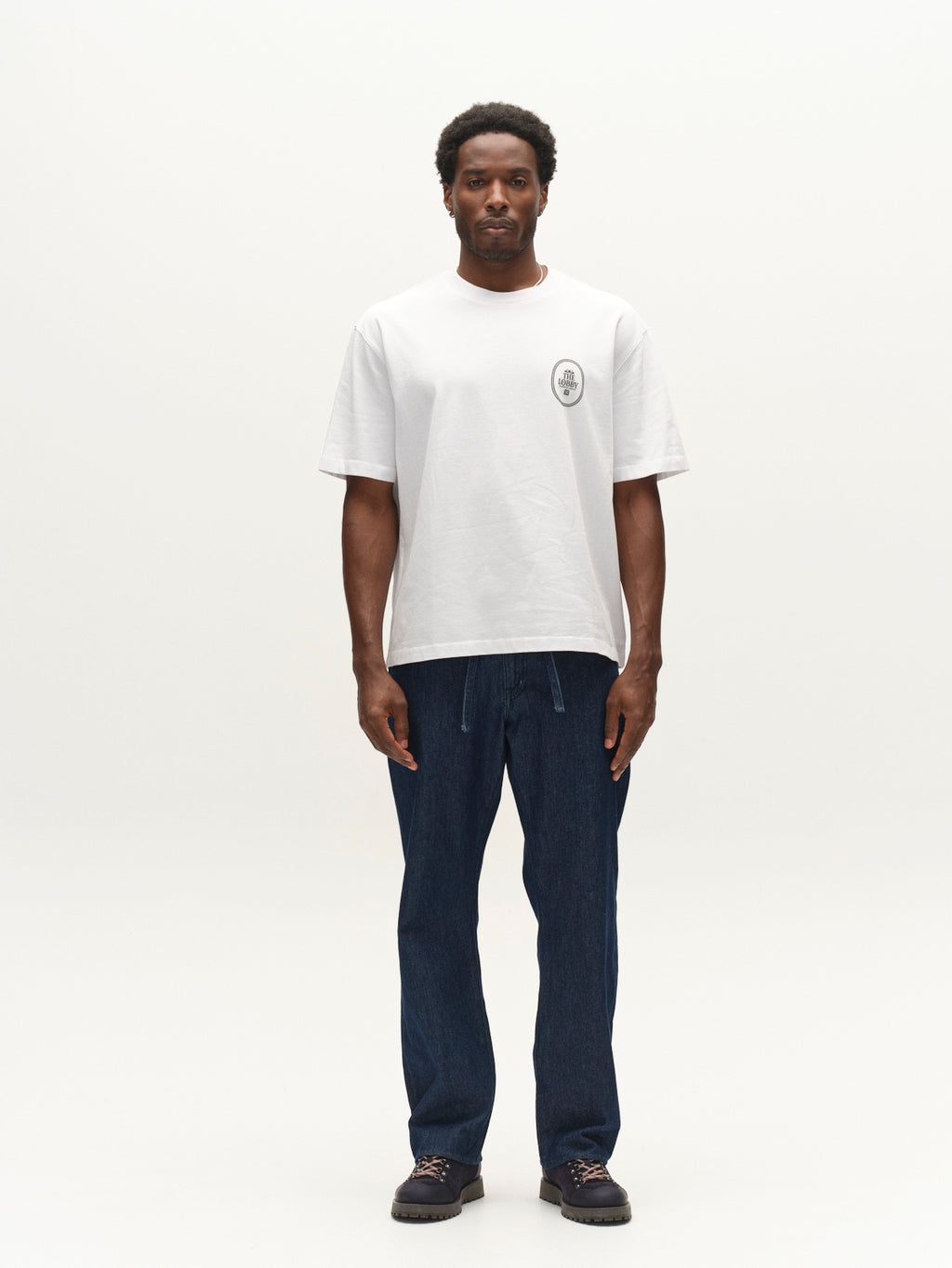 A man stands facing forward against a plain white background, wearing a white T-shirt crafted from an organic cotton blend with a small circular logo on the chest, Gabba GABnoah Tona Rinse Jeans in dark blue denim with a loose straight fit, and dark shoes.