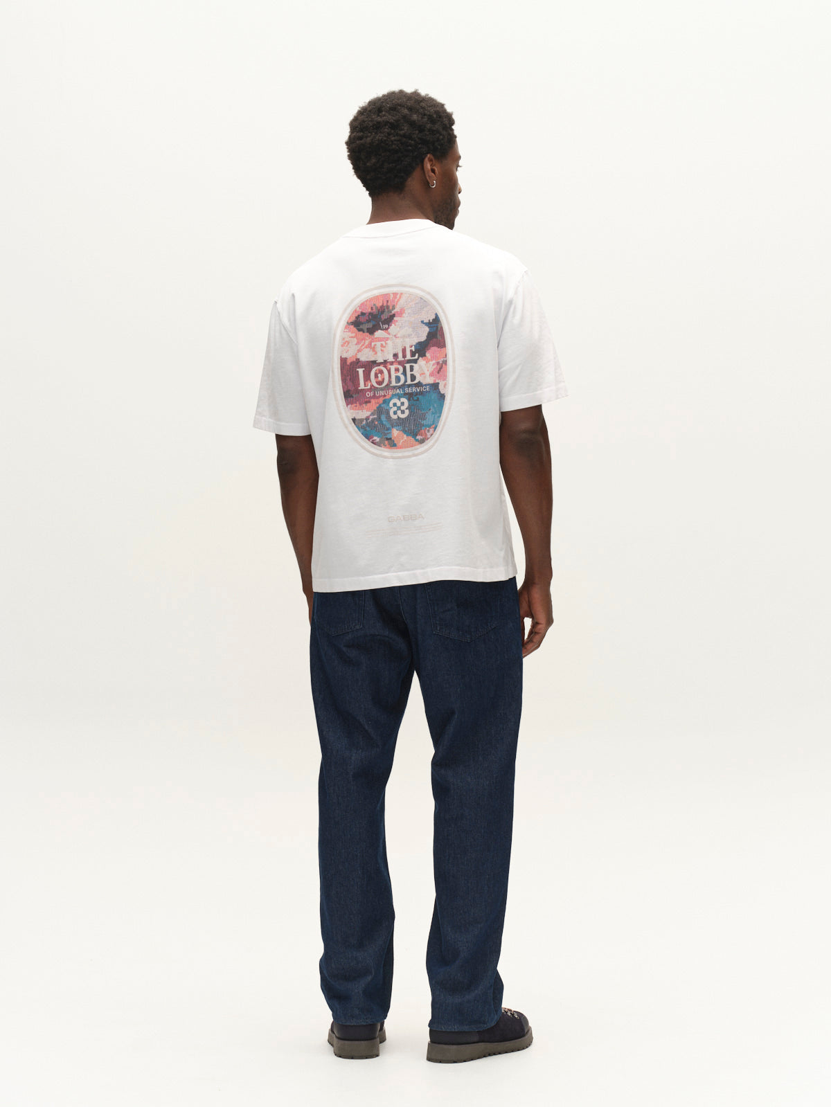 A man stands facing away, wearing a white T-shirt with a colorful graphic and the word LOBBY on the back, GABnoah Tona Rinse Jeans in dark blue denim by Gabba crafted from an organic cotton blend with a loose straight fit, and black shoes against a plain white background.
