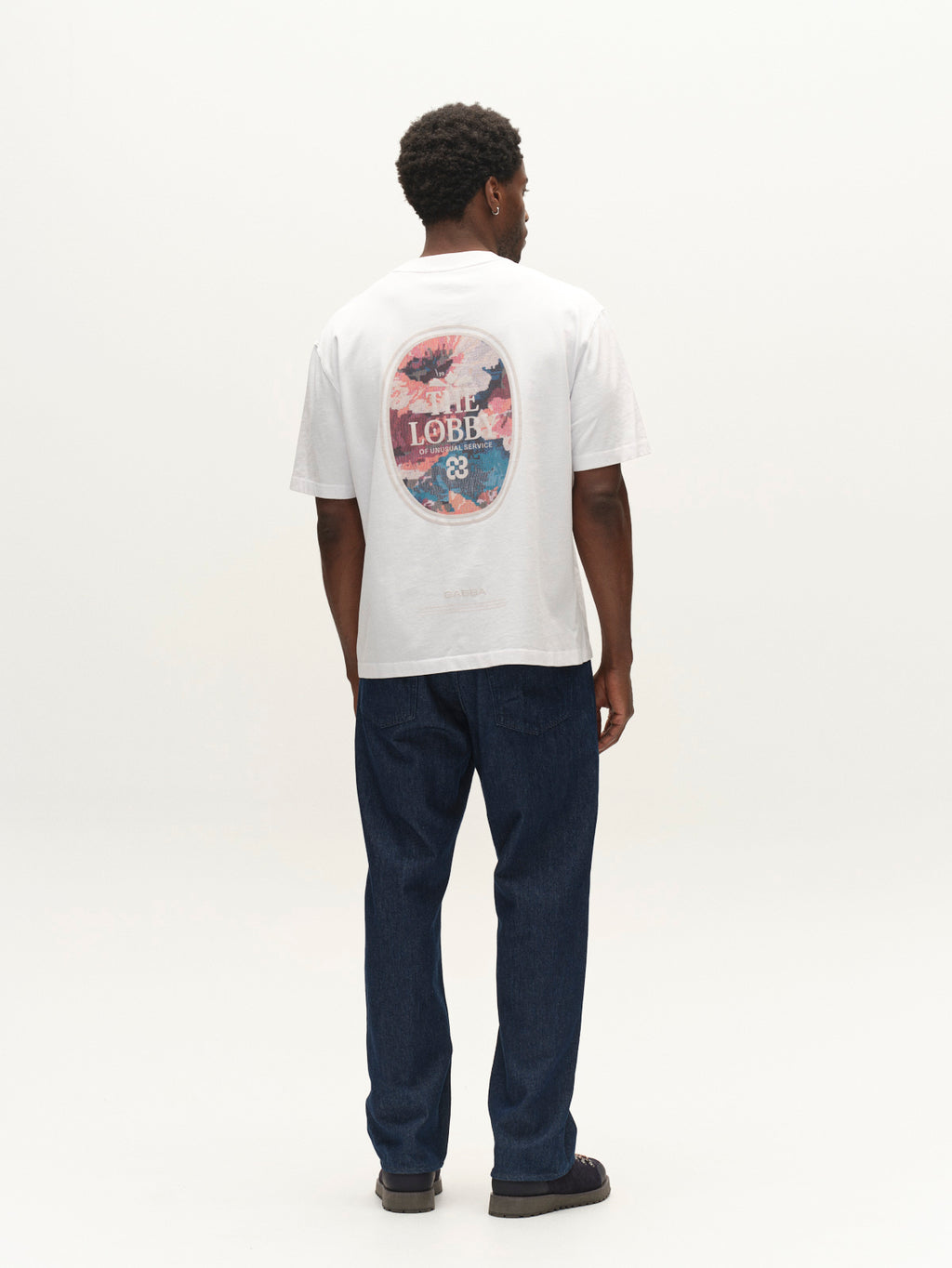 A man stands facing away, wearing a white T-shirt with a colorful graphic and the word LOBBY on the back, GABnoah Tona Rinse Jeans in dark blue denim by Gabba crafted from an organic cotton blend with a loose straight fit, and black shoes against a plain white background.