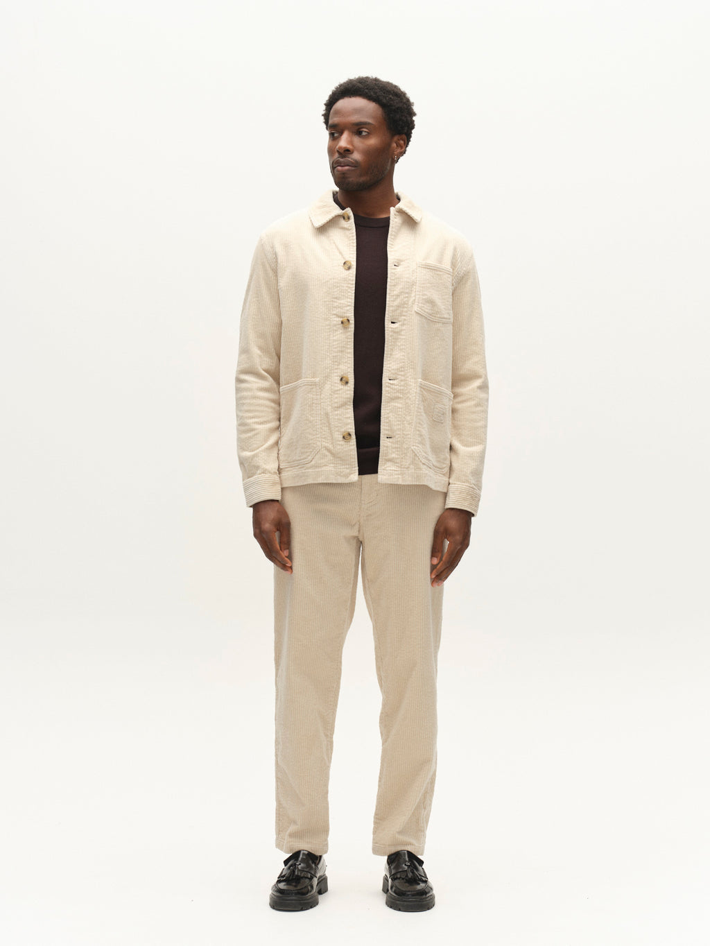 A man stands against a plain white background wearing the Gabba GABjoe Nopleats Ibre Pant in Birch, featuring a tapered fit and classic chino-inspired corduroy construction. He pairs the pants with a beige button-up jacket, a dark shirt underneath, and black shoes. He is looking slightly to his left with his arms relaxed at his sides.