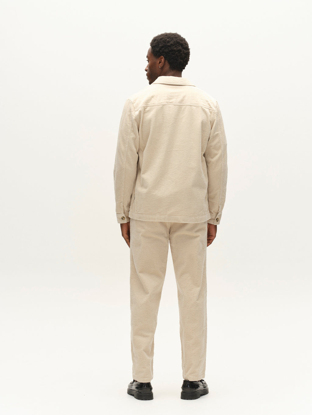 A person stands facing away in a plain, white studio setting, wearing the Gabba GABjoe Nopleats Ibre Pant in Birch—a light beige, tapered fit corduroy pant with classic chino construction—paired with a matching jacket and black shoes.