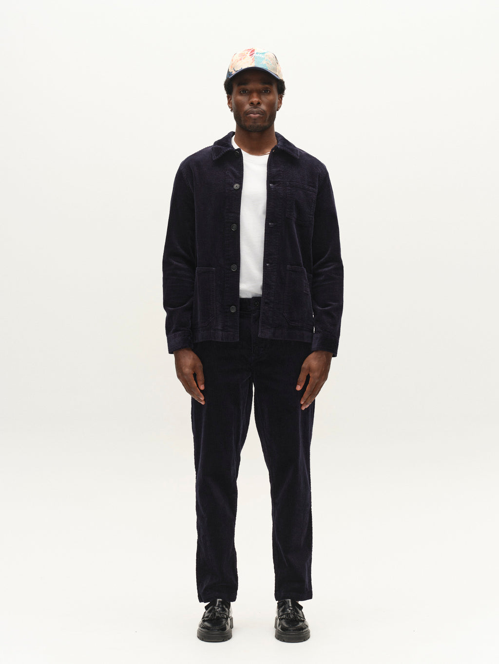 A man stands facing forward against a plain white background, wearing the Gabba GABjoe Nopleats Ibre Pant in Dark Navy—a pair of corduroy pants with classic chino construction and a tapered fit—paired with a dark jacket, white t-shirt, black shoes, and a colorful cap.