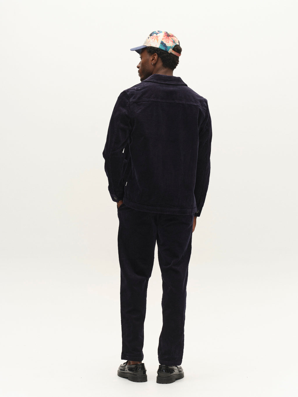 A man stands with his back to the camera against a plain white background, wearing Gabba’s GABjoe Nopleats Ibre Pant in Dark Navy, paired with a dark corduroy jacket, black shoes, and a colorful floral cap.
