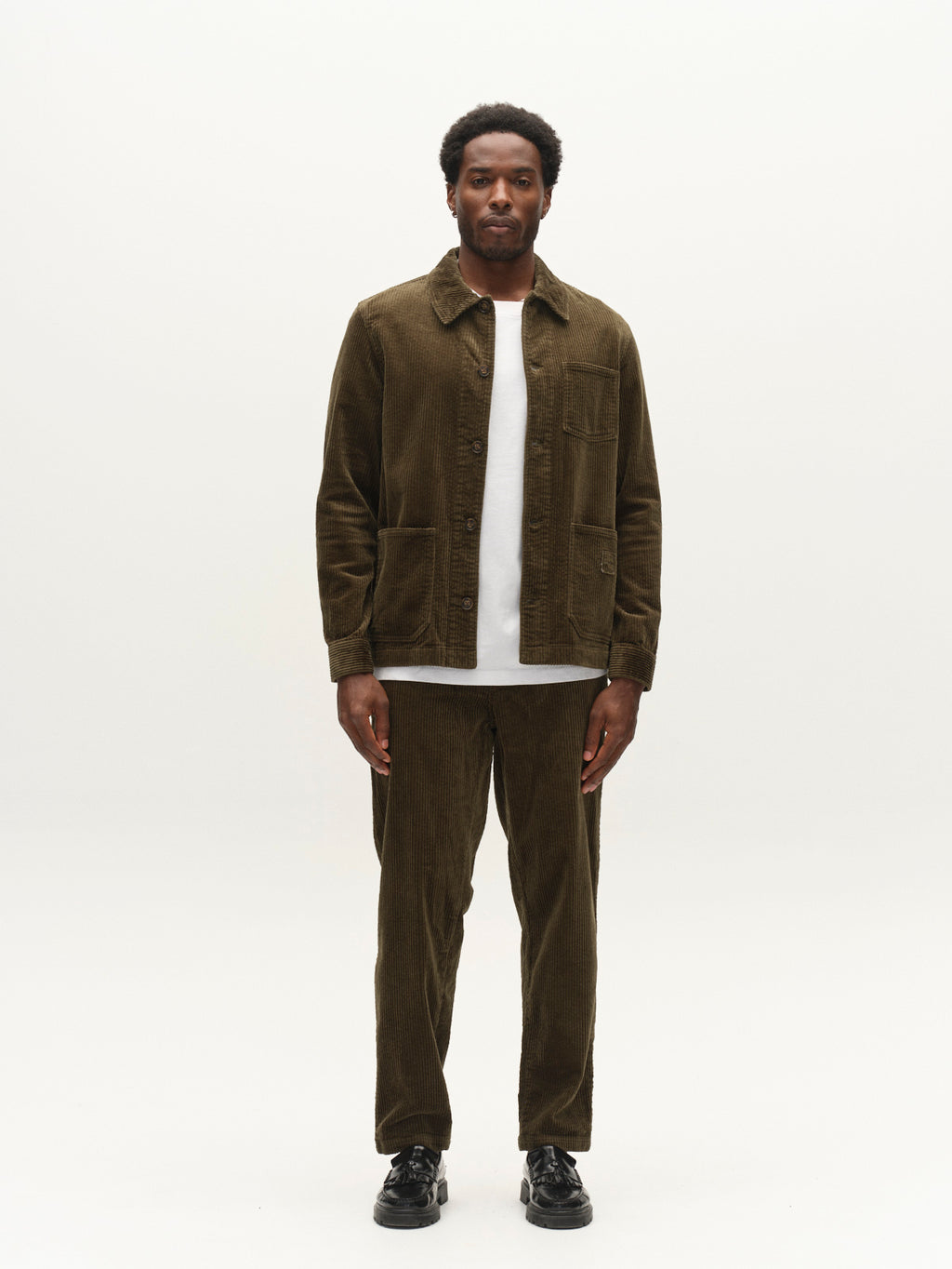A man stands facing forward against a plain white background, wearing Gabba’s GABjoe Nopleats Ibre Pant in Forest Night—a tapered fit corduroy design—paired with a matching olive green stretch corduroy jacket over a white shirt and black shoes.