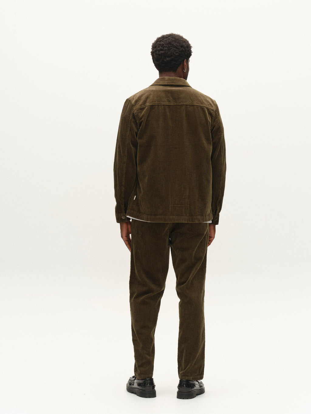 A person stands facing away from the camera, wearing the Gabba GABjoe Nopleats Ibre Pant in Forest Night, paired with a brown stretch corduroy jacket and black shoes, against a plain white background.
