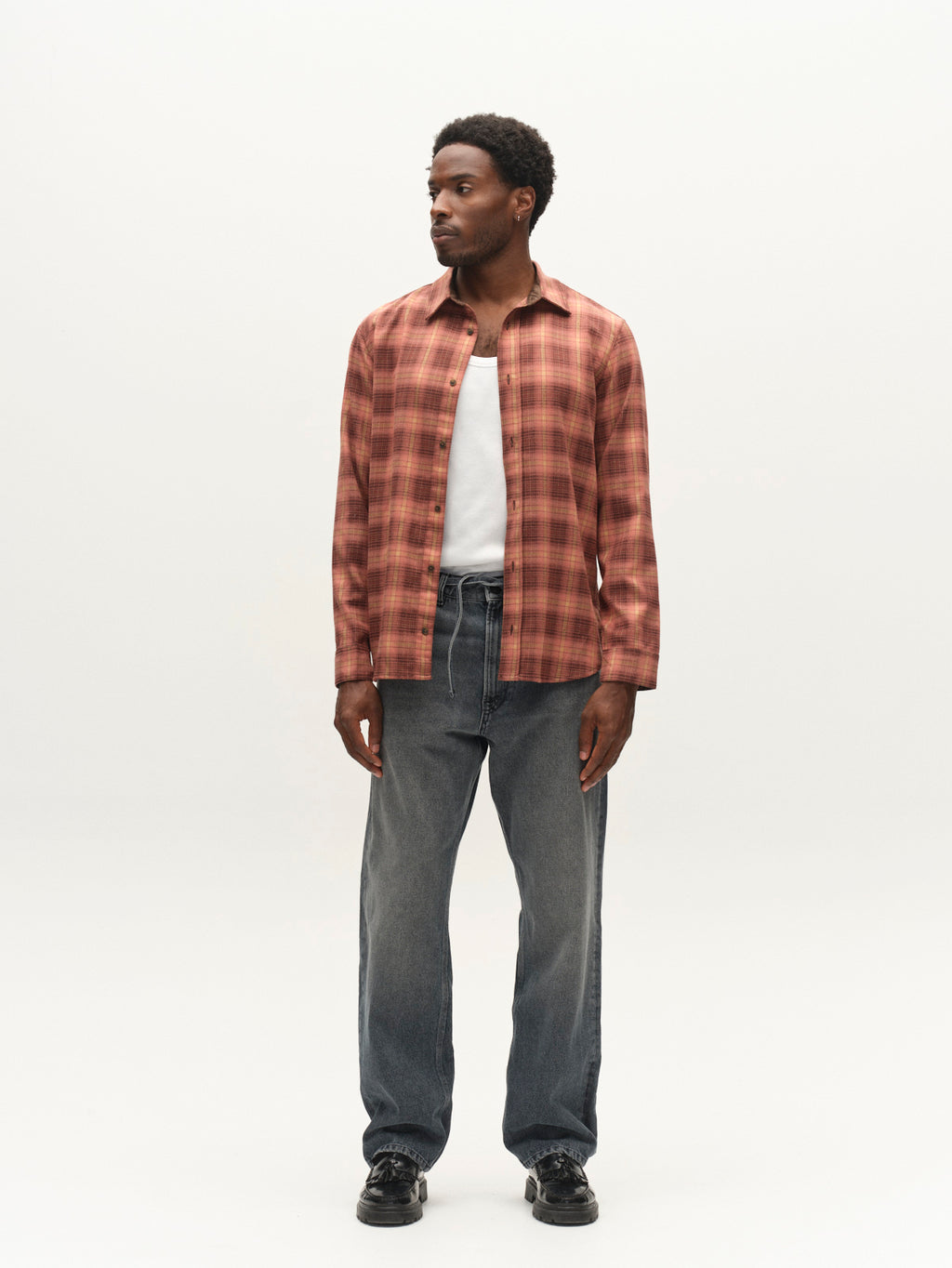 A man stands against a plain background wearing a red plaid shirt over a white T-shirt, Gabba GABnoah Primesto WW0030 Jeans in grey denim, and black shoes, looking to his left.