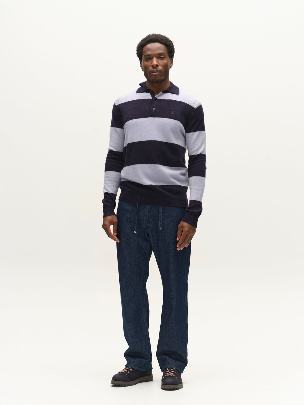 A man stands against a plain white background, wearing the Gabba GABworren Polo Stripes Aico LS Knit in navy stripe—a men’s regular fit long sleeve striped knit sweater—paired with blue pants and dark shoes.