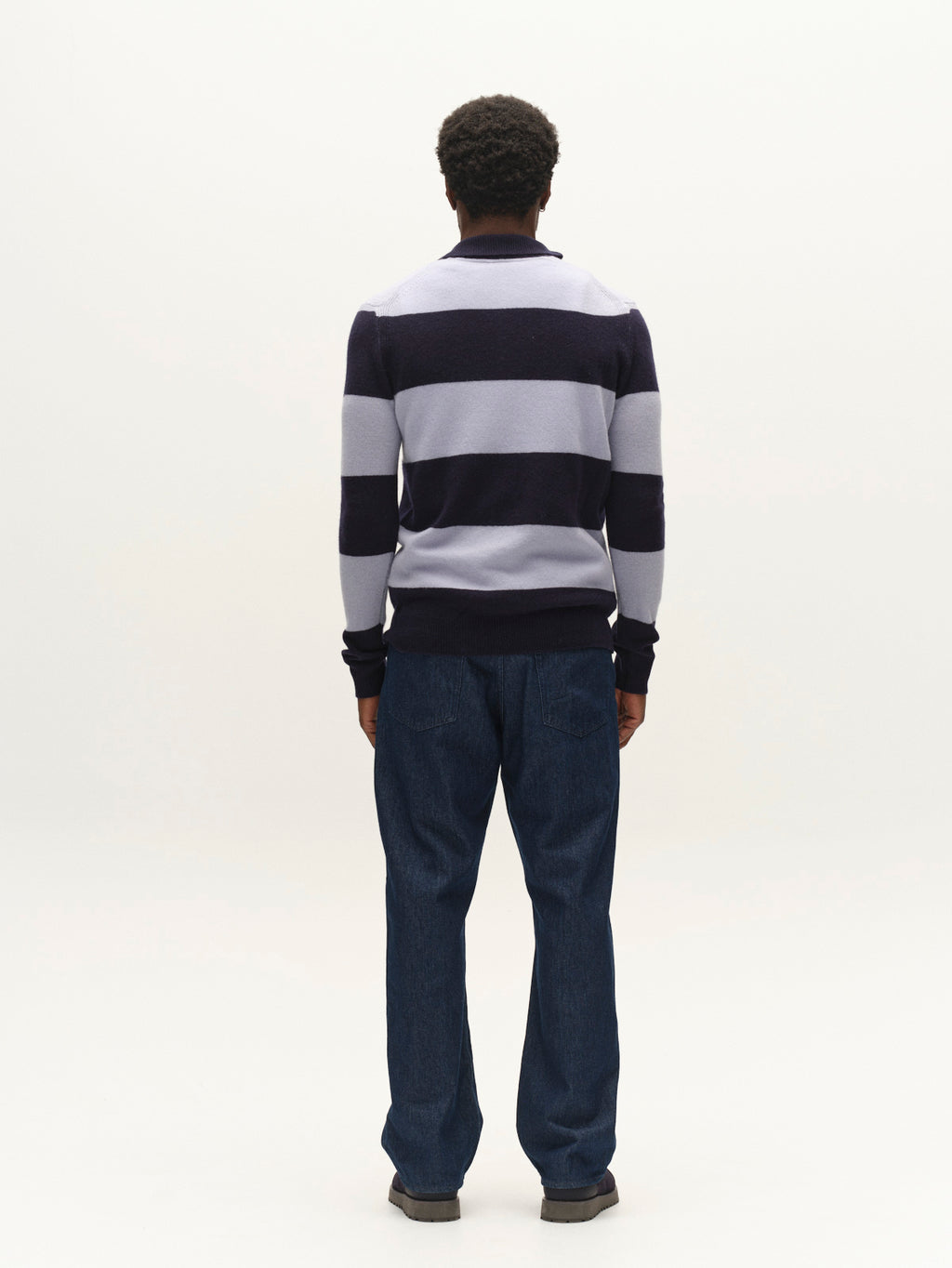 A person stands facing away from the camera, wearing the Gabba GABworren Polo Stripes Aico LS Knit in Navy Stripe, paired with blue jeans and gray shoes, against a plain white background.