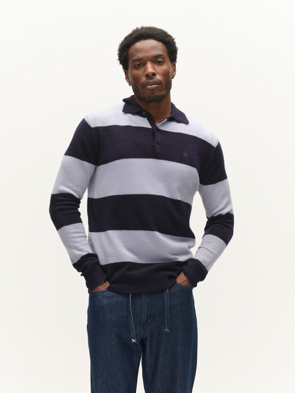 A man stands against a plain white background, wearing the Gabba GABworren Polo Stripes Aico LS Knit in Navy Stripe and blue jeans, with his hands in his pockets.
