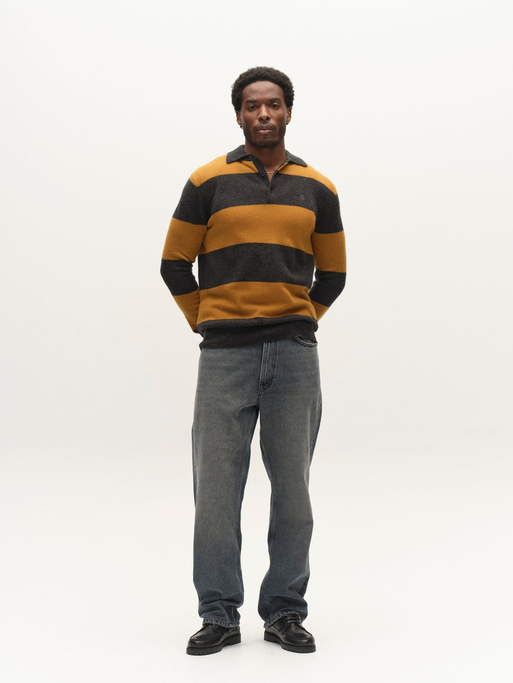 Against a plain white background, a man stands with his hands calmly behind his back, wearing the Gabba GABworren Polo Stripes Aico LS Knit in yellow stripes—styled in a classic men’s regular fit—paired with loose-fitting blue jeans and black shoes.