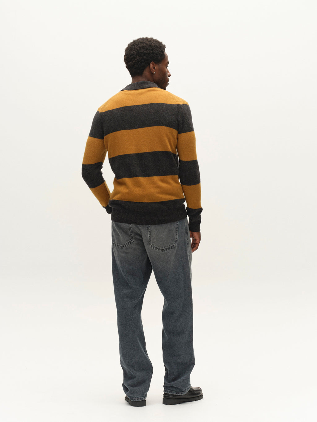 A person stands facing away against a plain white background, wearing Gabba’s GABworren Polo Stripes Aico LS Knit in yellow stripes and loose-fitting blue jeans.