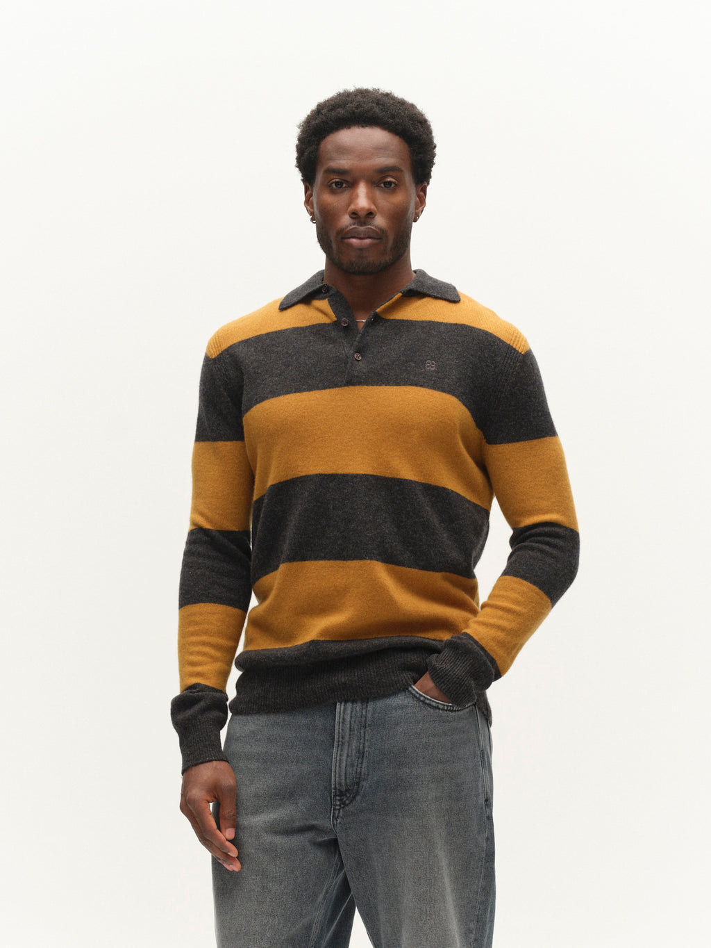 A man stands against a plain white background, wearing the Gabba GABworren Polo Stripes Aico LS Knit in yellow stripes—a soft wool knit sweater with mustard yellow and dark gray stripes—paired with medium-wash jeans. He looks directly at the camera with a neutral expression, highlighting the men’s regular fit of his outfit.