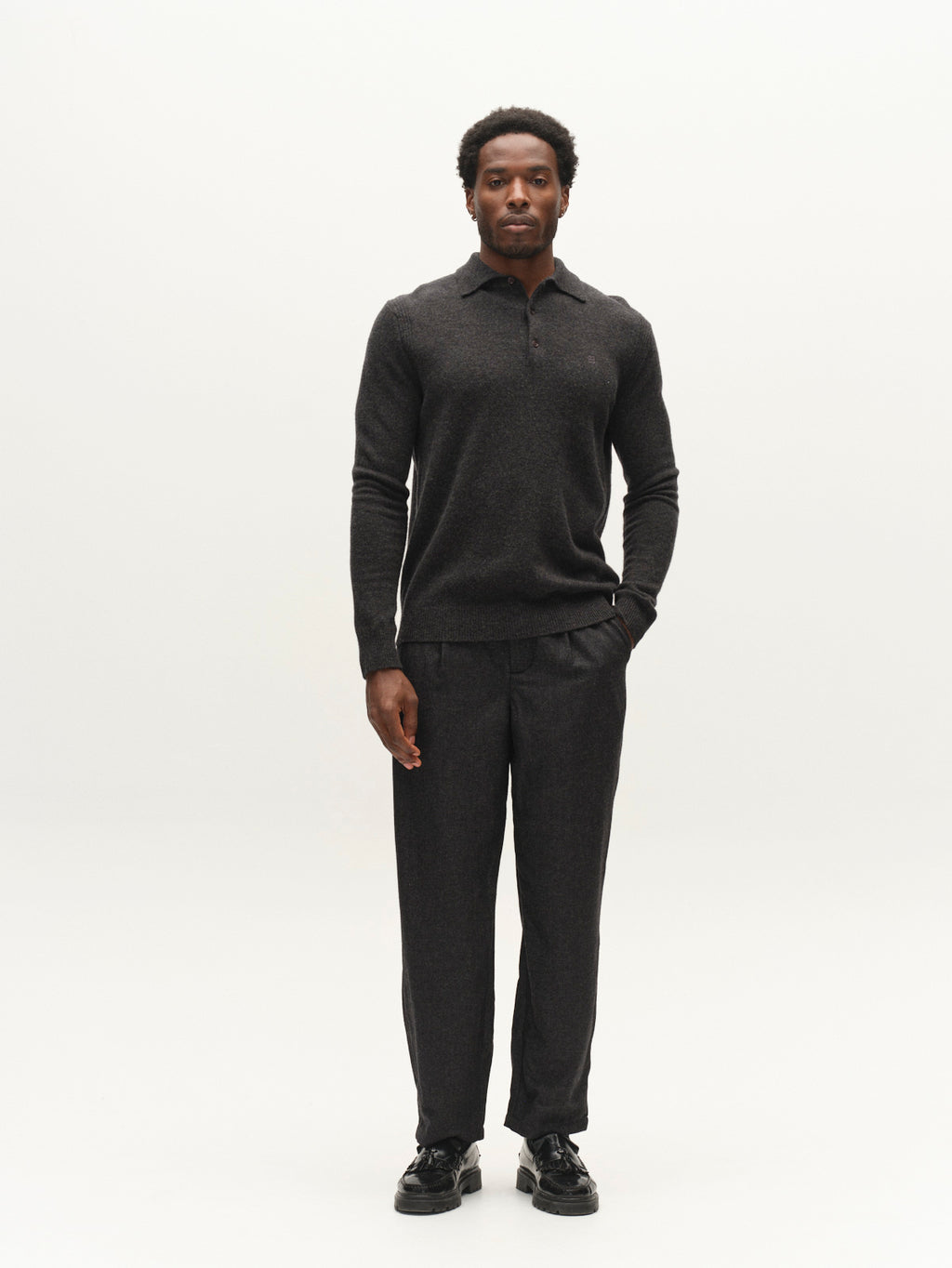 A man stands against a plain white background, wearing the Gabba GABworren Polo Aico LS Knit in Dark Grey Mel—a men’s regular fit sweater crafted from soft wool knit with a ribbed polo collar—paired with matching pants and black shoes, one hand in his pocket.
