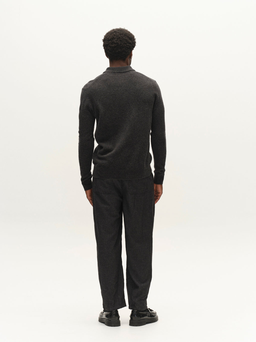 A person with short curly hair, dressed in the Gabba GABworren Polo Aico LS Knit sweater in Dark Grey Mel—a men’s regular fit style crafted from soft wool with a ribbed polo collar—paired with dark pants and black shoes, stands facing away from the camera against a plain white background.