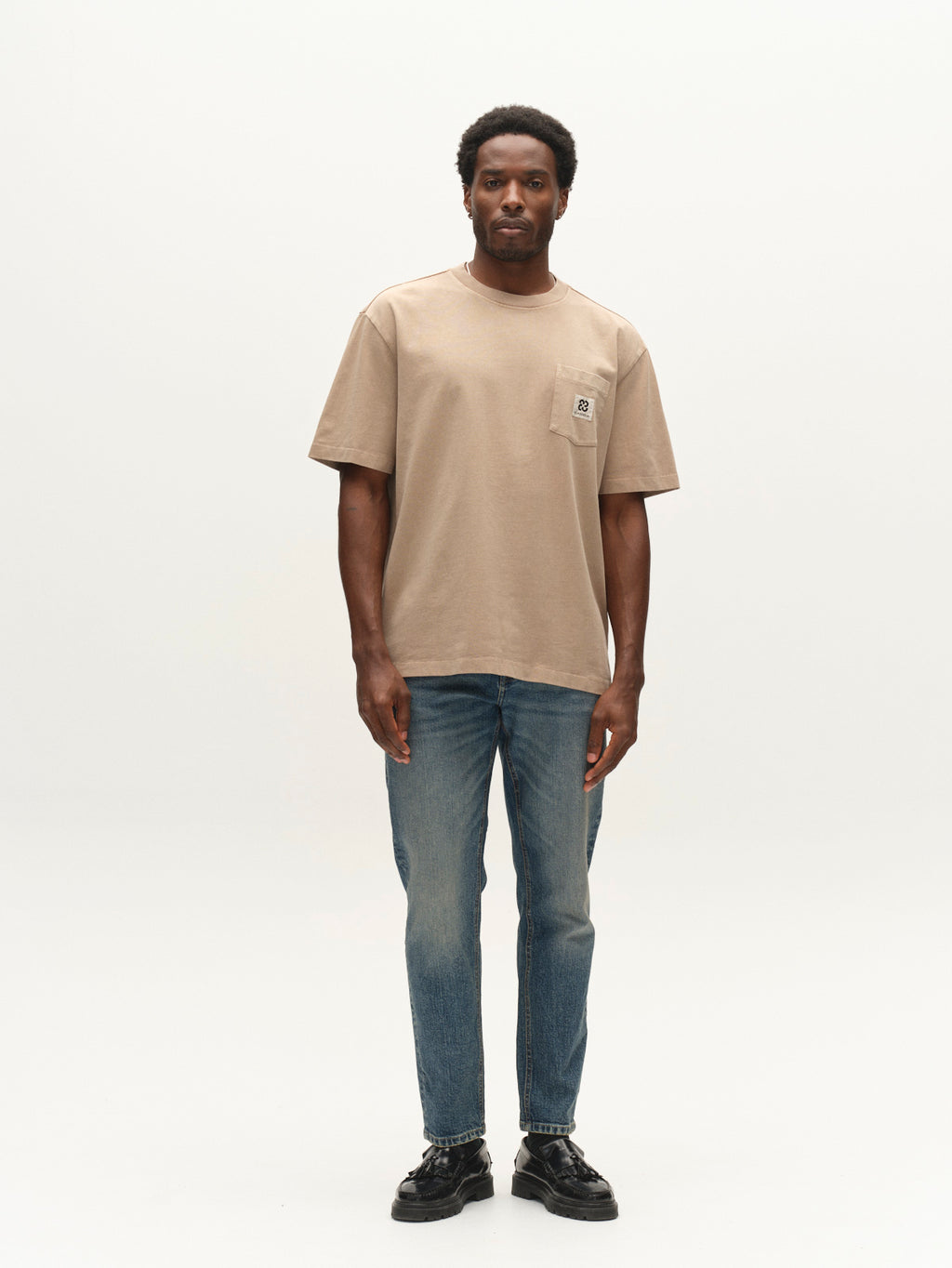 A man stands against a plain white background wearing the Gabba GABnigel Wix Pkt Boxy Hour SS T-Shirt in Sand Crocker—a boxy heavyweight cotton tee with a chest pocket—paired with blue jeans and black shoes.