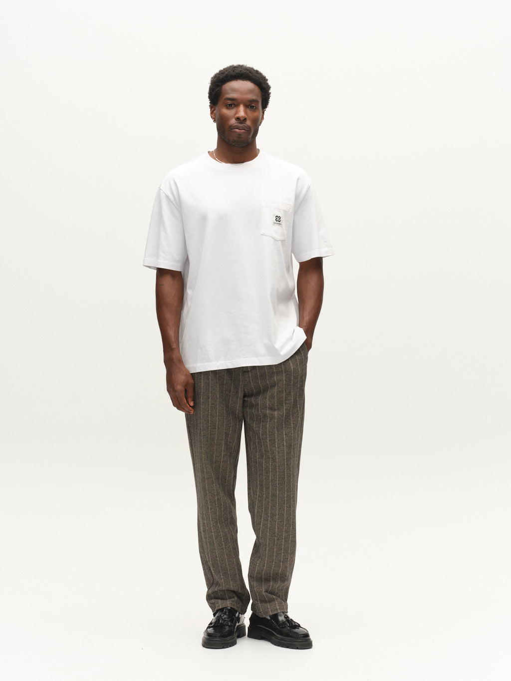 A man stands against a plain white background, wearing the Gabba GABnigel Wix Pkt Boxy Hour SS T-Shirt in white, crafted from heavyweight cotton with a chest pocket, paired with gray pinstriped pants and black shoes.
