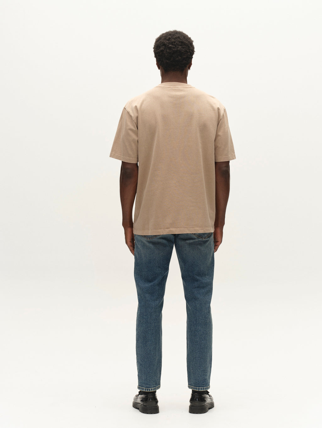 A person stands facing away from the camera, wearing the Gabba GABnigel Wix Pkt Boxy Hour SS T-Shirt in Sand Crocker, paired with blue jeans and black shoes, against a plain light background.