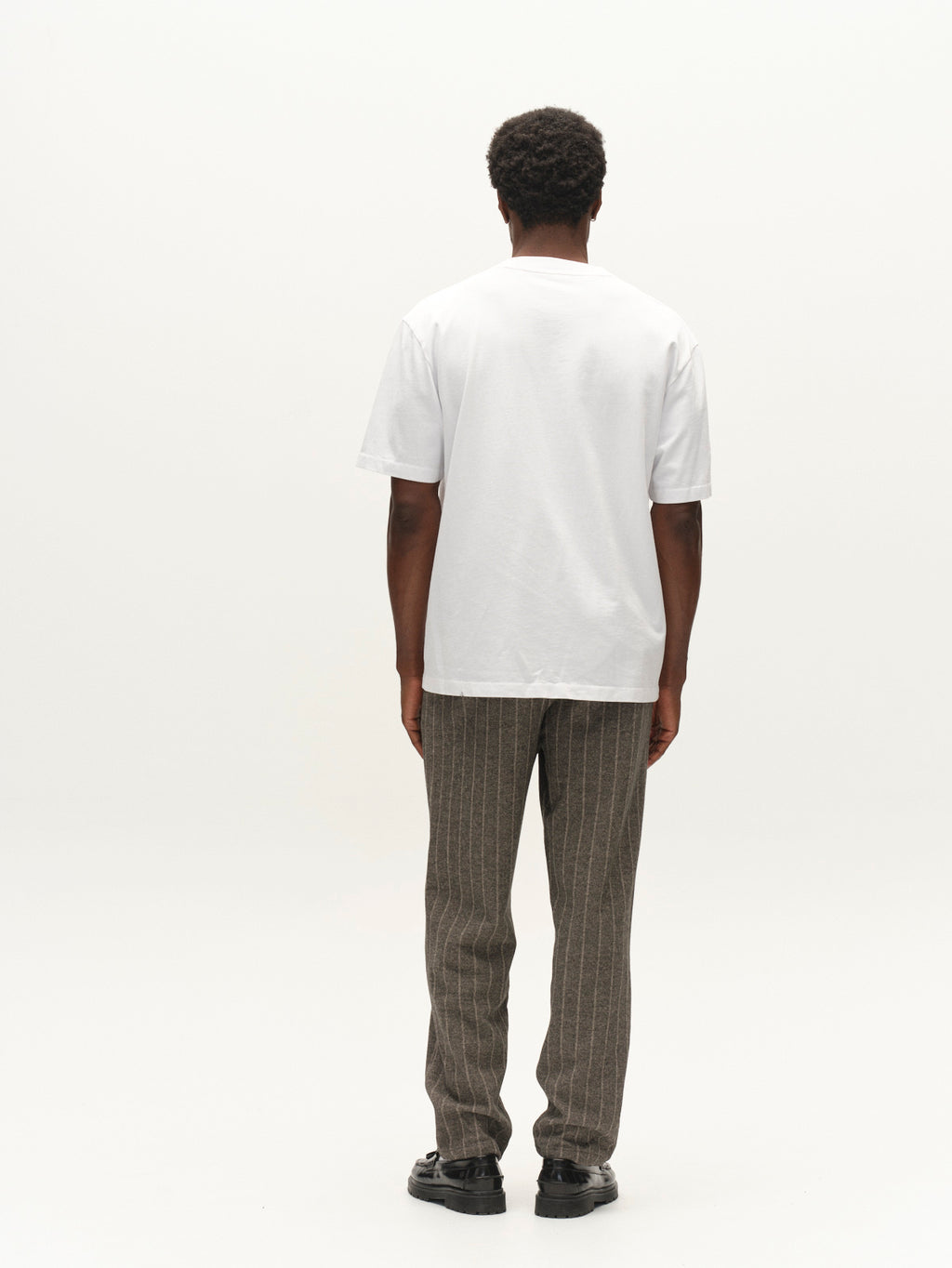 A person stands facing away, wearing the Gabba GABnigel Wix Pkt Boxy Hour SS T-Shirt in white, paired with brown pinstripe pants and black shoes against a plain white background.