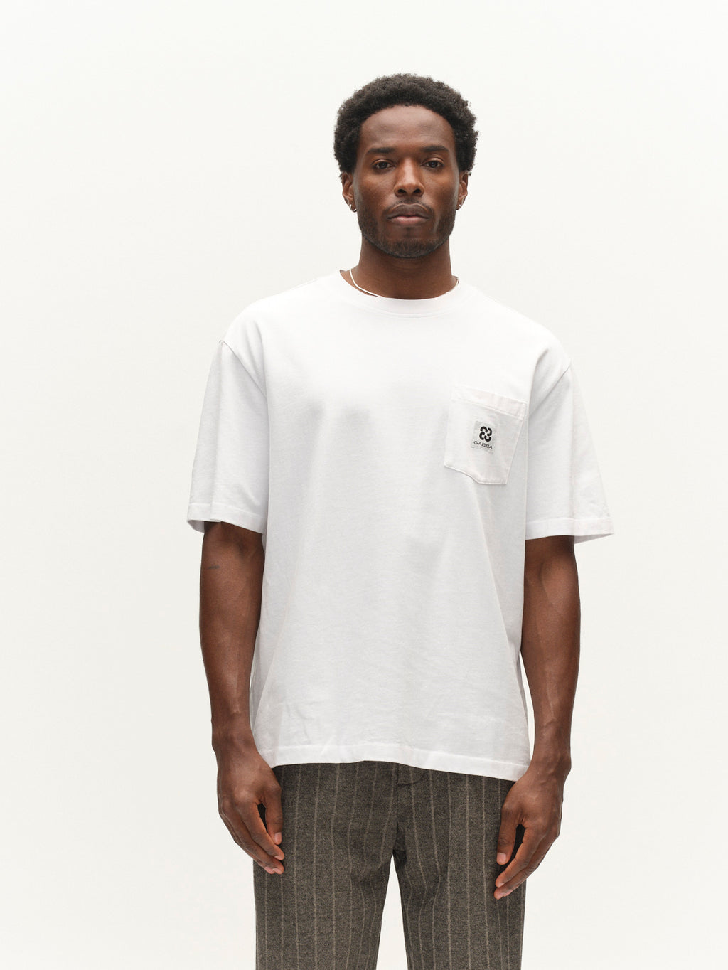 A man wearing the Gabba GABnigel Wix Pkt Boxy Hour SS T-Shirt in white—a plain, heavyweight cotton tee with a boxy fit, small logo on the chest pocket—paired with gray pinstriped trousers, stands against a white background.