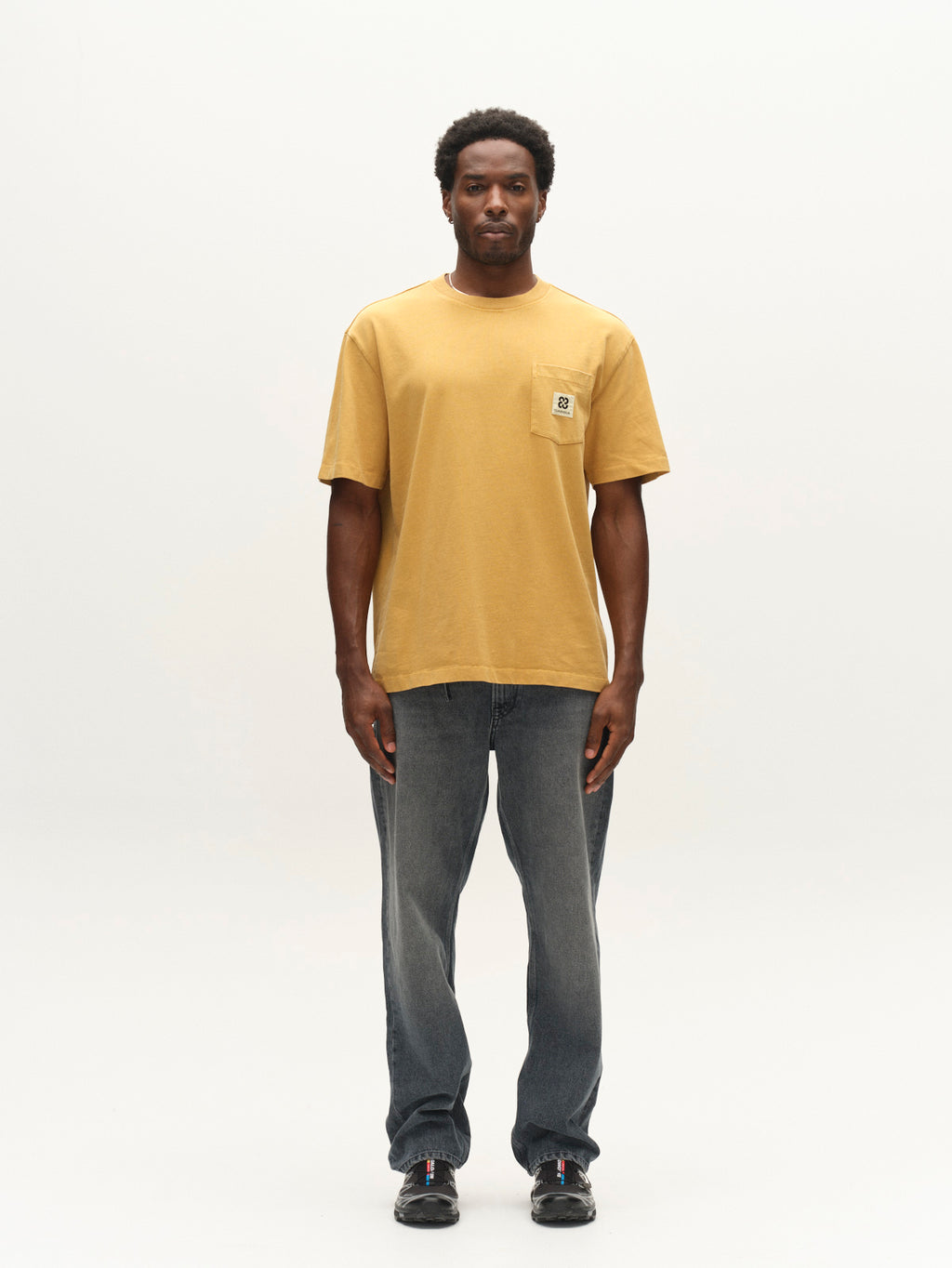 A man stands facing forward against a plain white background, wearing a Gabba GABnigel Wix Pkt Boxy Hour SS T-Shirt in mustard yellow with a front pocket, paired with blue jeans and black shoes.