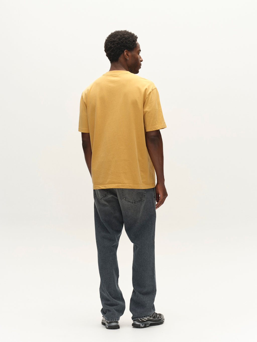 A man stands facing away from the camera against a plain light background, wearing the Gabba GABnigel Wix Pkt Boxy Hour SS T-Shirt in Mustard Yellow—a boxy, heavyweight cotton tee—paired with dark jeans and sneakers. The subtle Gabba brand mark appears on the back of his T-shirt.