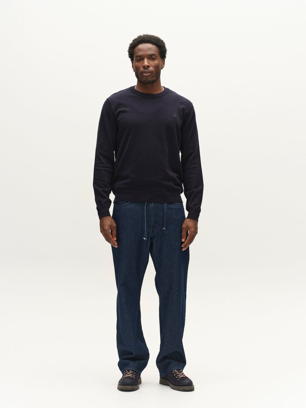 A man wearing a navy GABfumo Element Spy LS Knit sweater, dark blue trousers with a drawstring waist, and dark hiking-style boots.