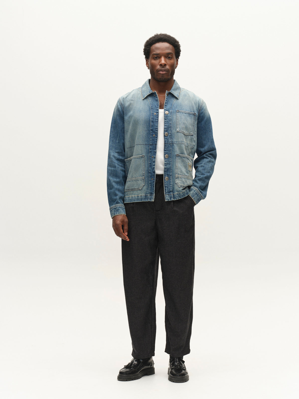 A man stands facing forward, wearing the Gabba GABbeta Wolv DS0004 LS Overshirt in dark blue denim over a white shirt, paired with loose dark trousers and black shoes, set against a plain, light background. The look channels workwear inspiration with an effortlessly modern edge.